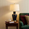 29" Navy Blue And Gold Urn Bedside Lamp With Drum Shade styled in a furnished interior with a cream wall on a black floor.