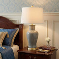 29" Gray And Gold Urn Bedside Lamp With Drum Shade styled in a furnished interior with a light gray wall on a black floor.