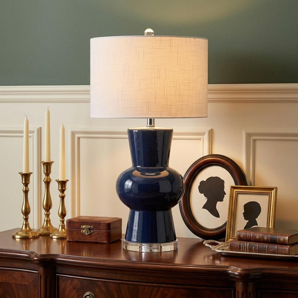 29" Clear Novelty Table Lamp With Drum Shade displayed on a dark wood tabletop in a furnished interior against a gray wall.