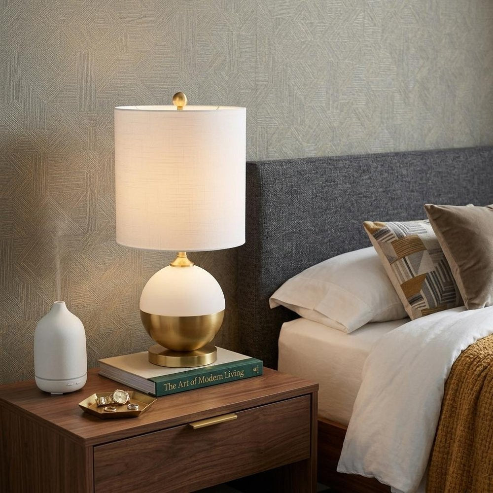 24" Gold Globe Table Lamp With Drum Shade displayed on a dark wood tabletop in a furnished interior against a tan wall.
