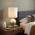 24" Gold Globe Table Lamp With Drum Shade displayed on a dark wood tabletop in a furnished interior against a tan wall.