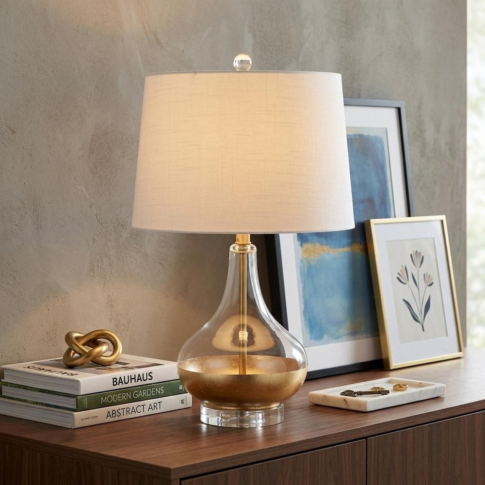 25" Clear Novelty Table Lamp With Drum Shade displayed on a dark wood tabletop in a furnished interior against a tan wall.