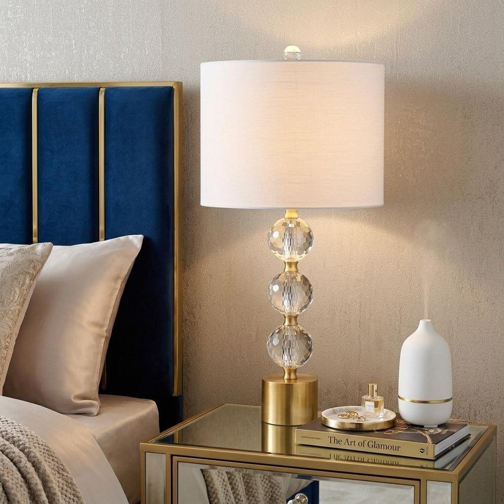 25" Gold Novelty Table Lamp With Drum Shade displayed on a dark wood tabletop in a furnished interior against a cream wall.