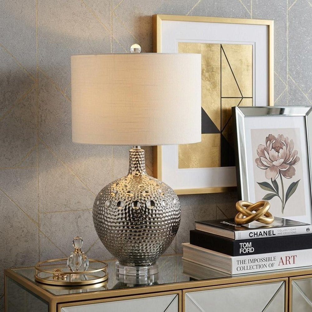 27" Clear Novelty Bedside Lamp With Drum Shade styled in a furnished interior with a tan wall on a tiled floor.