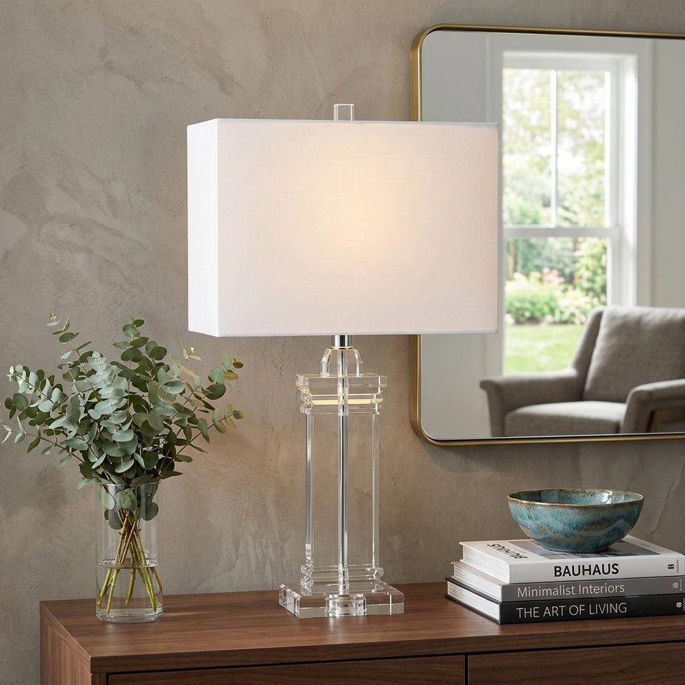25" Clear Novelty Table Lamp With Shade displayed on a dark wood tabletop in a furnished interior against a tan wall.