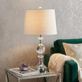 26" Silver Urn Bedside Lamp With Empire Shade styled in a furnished interior with a light gray wall on a dark wood floor.