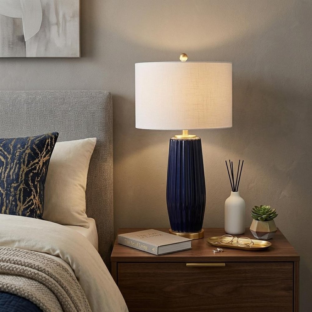 29" Navy Blue And Gold Table Lamp With Drum Shade displayed on a tabletop in a furnished interior against a tan wall.
