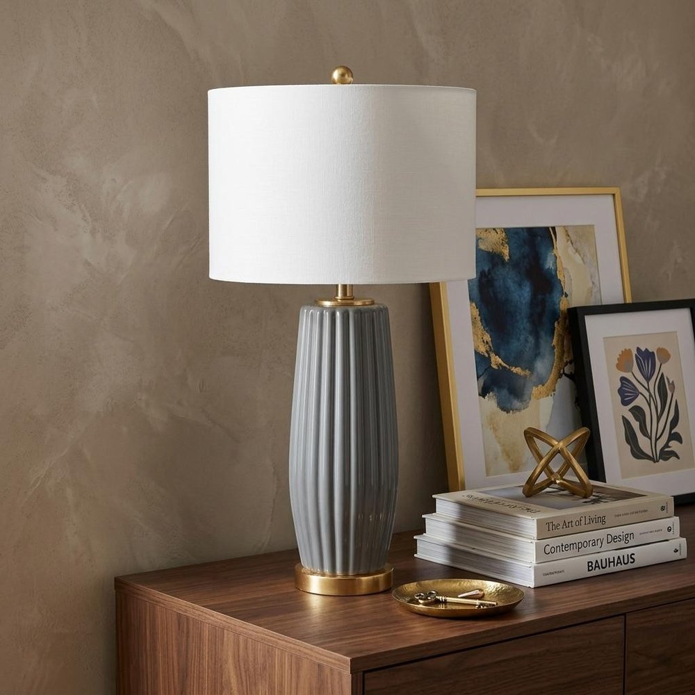 29" Dark Gray And Gold Table Lamp With Drum Shade displayed on a dark wood tabletop in a furnished interior against a brown wall.