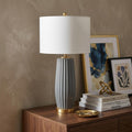 29" Dark Gray And Gold Table Lamp With Drum Shade displayed on a dark wood tabletop in a furnished interior against a brown wall.