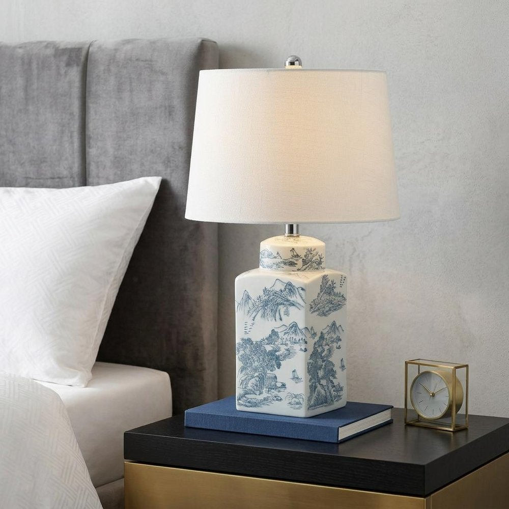 25" Blue And White Table Lamp With Drum Shade displayed on a tabletop in a furnished interior against a light gray wall.