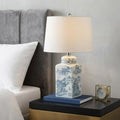 25" Blue And White Table Lamp With Drum Shade displayed on a tabletop in a furnished interior against a light gray wall.