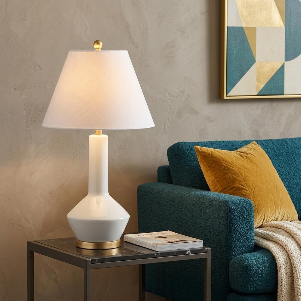 29" White And Gold Urn Table Lamp With Empire Shade displayed on a dark wood tabletop in a furnished interior against a tan wall.