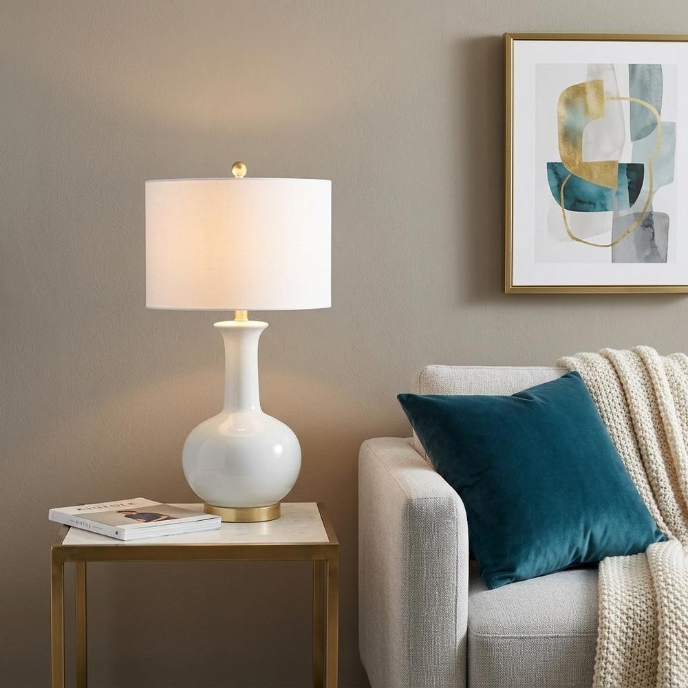 27" White And Gold Novelty Table Lamp With Drum Shade displayed on a dark wood tabletop in a furnished interior against a tan wall.