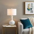 27" White And Gold Novelty Table Lamp With Drum Shade displayed on a dark wood tabletop in a furnished interior against a tan wall.