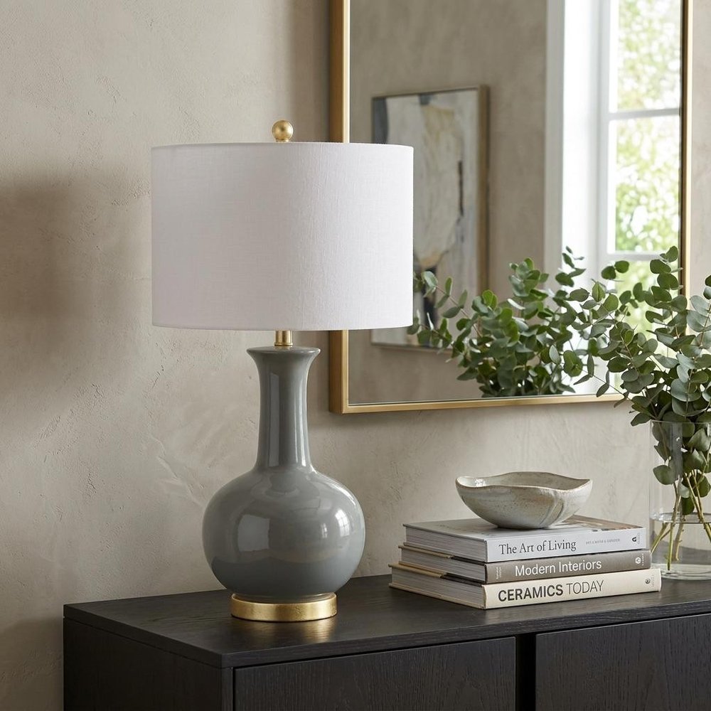 27" Steel Blue And Gold Novelty Table Lamp With Drum Shade displayed on a tabletop in a furnished interior against a tan wall.