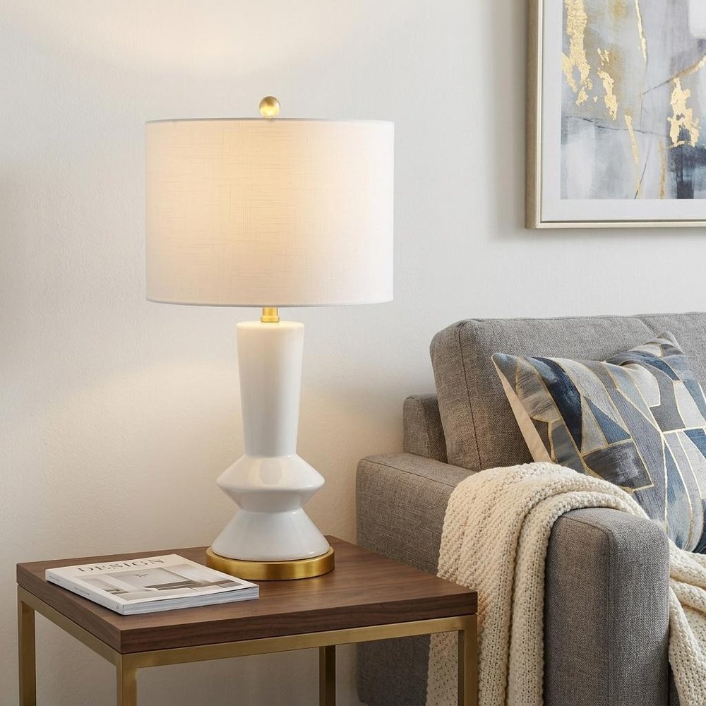 27" White And Gold Geometric Table Lamp With Drum Shade displayed on a dark wood tabletop in a furnished interior against an off-white wall.