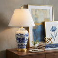27" Gold Novelty Table Lamp With Empire Shade displayed on a dark wood tabletop in a furnished interior against a tan wall.