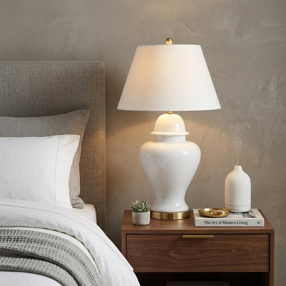 33" White And Gold Urn Table Lamp With Empire Shade displayed on a dark wood tabletop in a furnished interior against a tan wall.