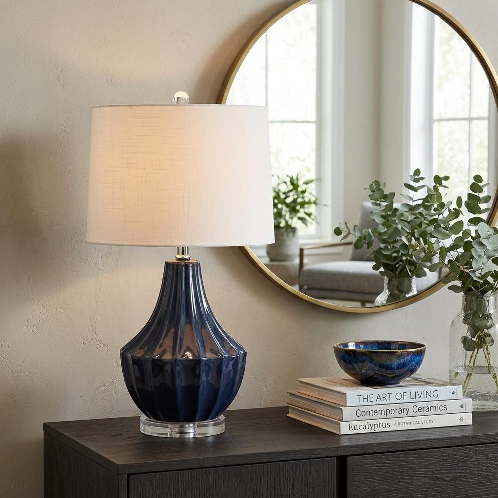 25" Navy Blue And Clear Novelty Table Lamp With Drum Shade displayed on a tabletop in a furnished interior against a tan wall.