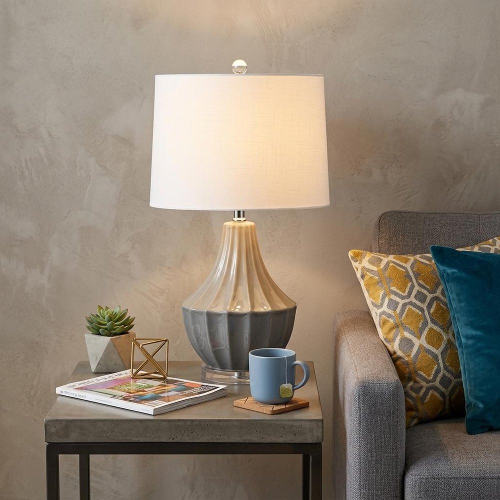 25" Gray And Clear Novelty Table Lamp With Drum Shade displayed on a dark wood tabletop in a furnished interior against a tan wall.