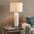 32" Silver Novelty Table Lamp With Drum Shade displayed on a dark wood tabletop in a furnished interior against a tan wall.
