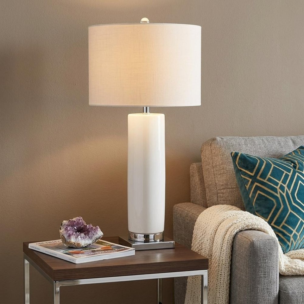 32" Silver Novelty Table Lamp With Drum Shade displayed on a dark wood tabletop in a furnished interior against a tan wall.