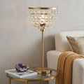 25" Gold Novelty Table Lamp With Drum displayed on a dark wood tabletop in a furnished interior against a light gray wall.