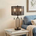 27" Antiqued Bronze Novelty Table Lamp With Drum Shade displayed on a wood tabletop in a furnished interior against a cream wall.