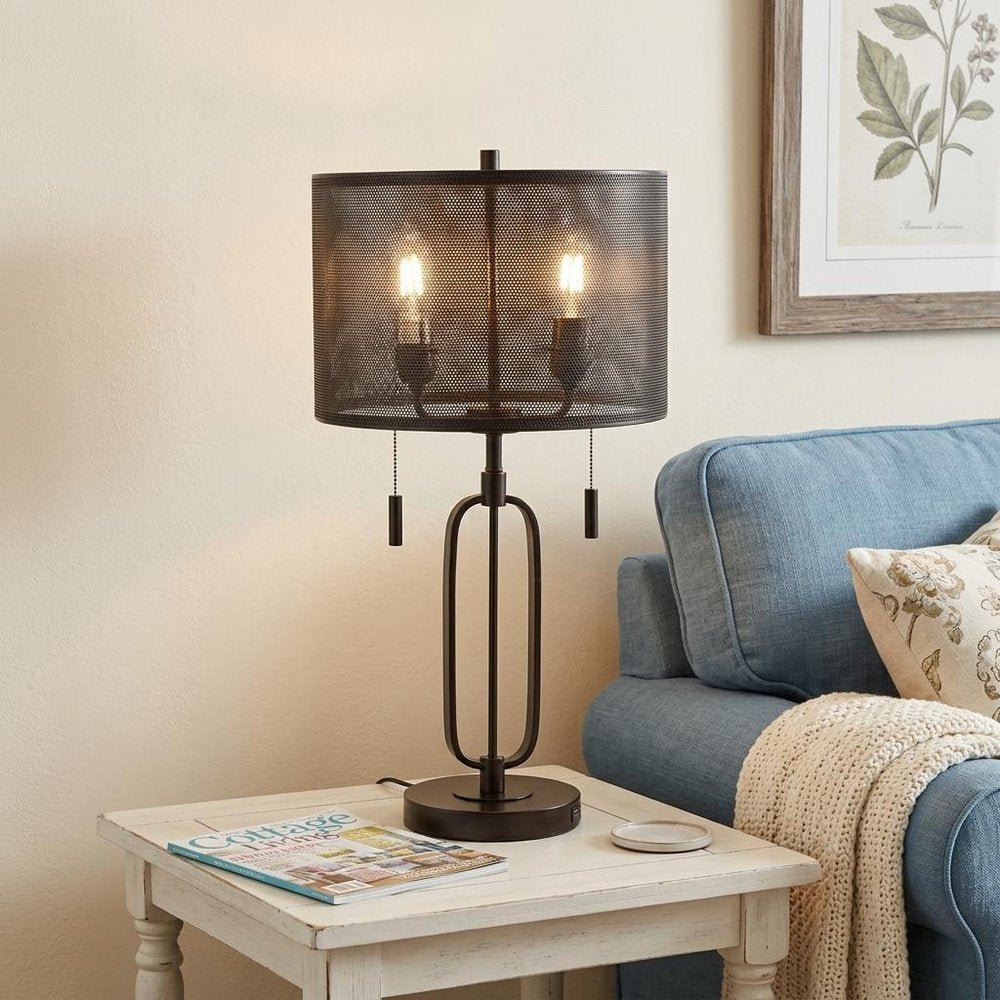 27" Antiqued Bronze Novelty Table Lamp With Drum Shade displayed on a wood tabletop in a furnished interior against a cream wall.