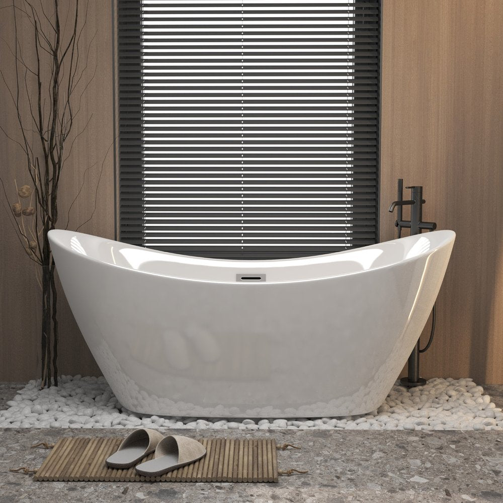 A white acrylic oval bathtub sits on white pebbles in front of a window with blinds, with a bamboo mat and slippers on the floor.