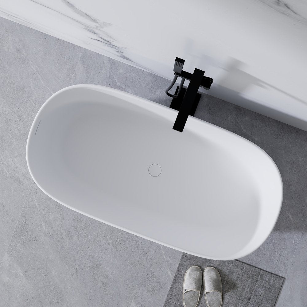 A white oval acrylic bathtub with a black faucet on a grey tiled floor with marble accent wall and slippers.