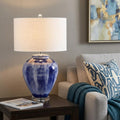 26" Blue And Clear Urn Table Lamp With Drum Shade displayed on a tabletop in a furnished interior against a tan wall.