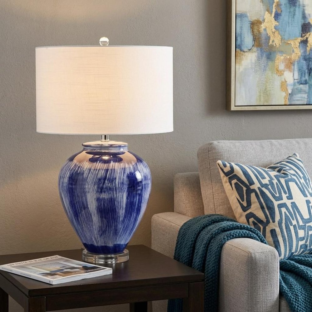 26" Blue And Clear Urn Table Lamp With Drum Shade displayed on a tabletop in a furnished interior against a tan wall.