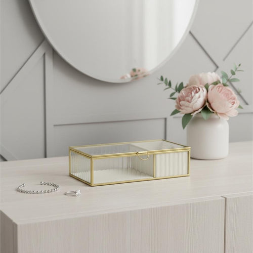 Rectangular gold glass jewelry box with two sections on a light wood dresser, with a bracelet and ring in front and a vase of pink peonies behind.
