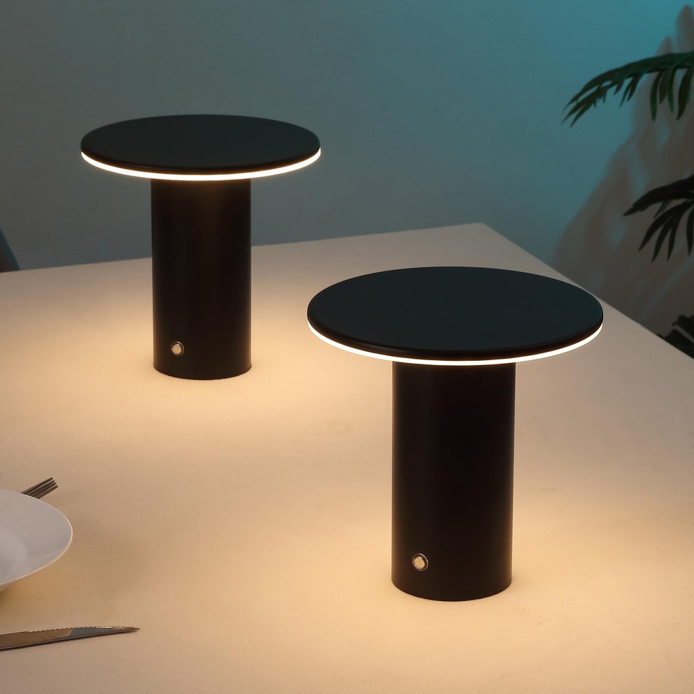 Two black cylindrical table lamps with round tops and white LED light rings on a light-colored table.