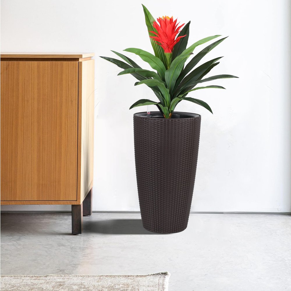 Tall brown woven self-watering planter with a red bromeliad plant next to a wooden cabinet.