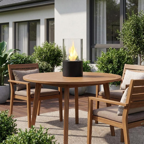 Round wooden patio table with chairs and a black and clear glass tabletop bioethanol firepit on a rug.