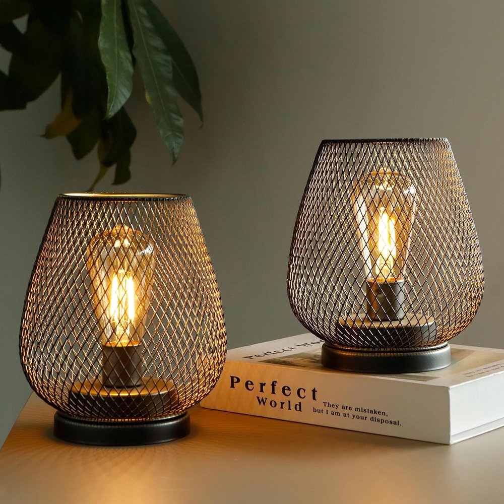 Two antiqued bronze pierced geometric cordless lantern lamps with Edison bulbs on a wooden surface with a book.