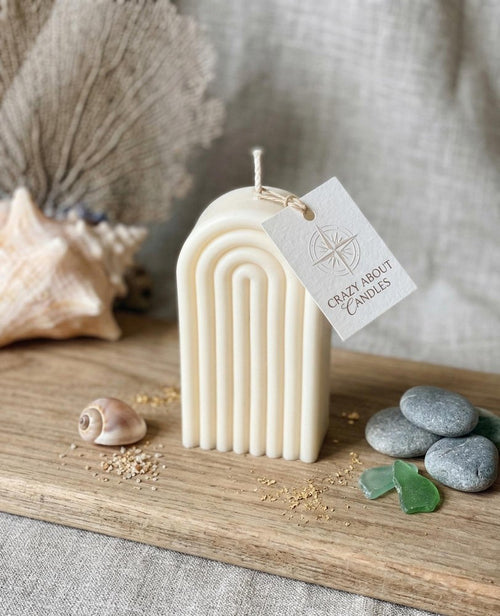 A white arched candle with a tag sits on a wooden board with seashells, sea glass, and pebbles.