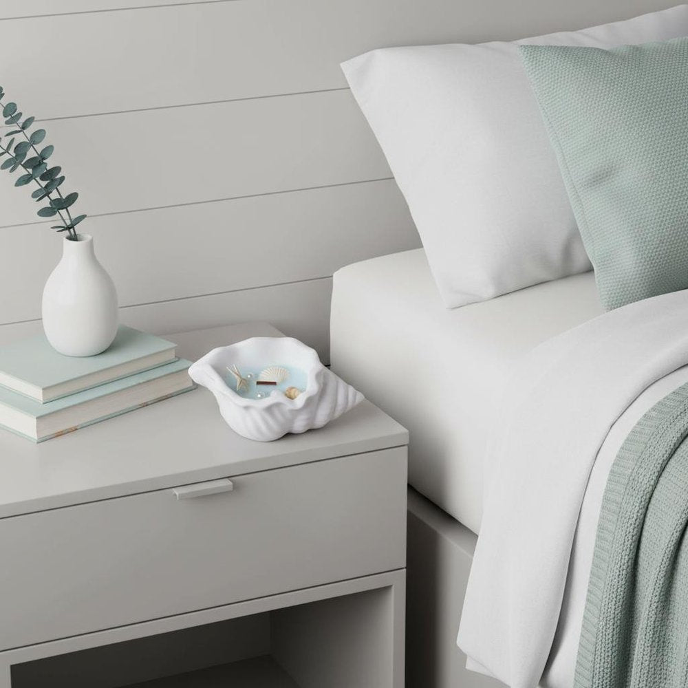 6" Light Blue Sea Salt And Orchid Scented Candle In White Resin Novelty Holder With Shells displayed on a tabletop in a furnished interior against a light gray wall.