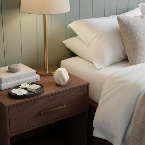 Ivory rose petals scented candle on a wooden nightstand next to a bed with white linens and a green paneled wall.