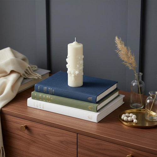 Ivory white carved pillar candle on a stack of books with a dried grass in a vase on a wooden dresser.