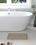 Beige gray teak non-slip bath mat with horizontal slats on a tiled bathroom floor next to a white bathtub.