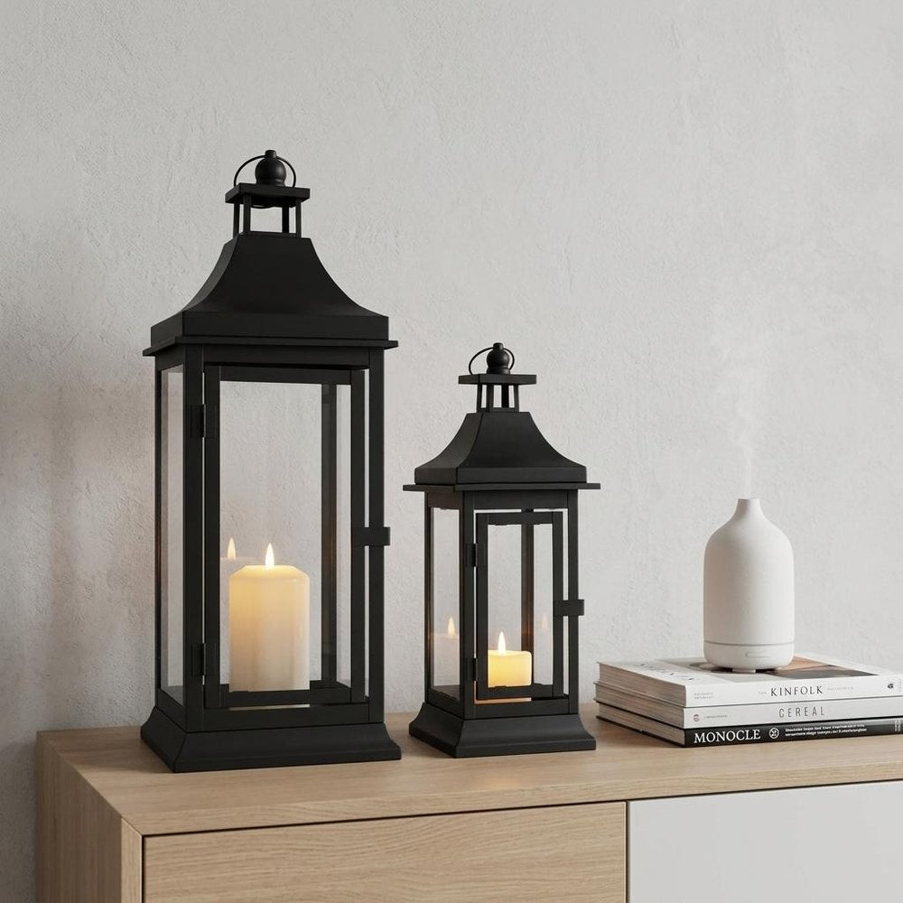 Two black metal and glass lanterns with lit candles sit on a wooden surface in front of a white wall.
