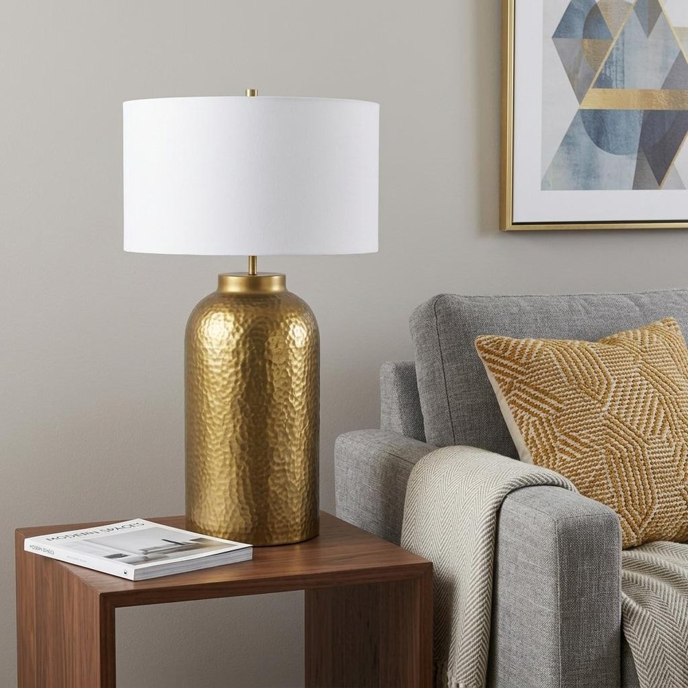 30" Gold Novelty Table Lamp With Drum Shade displayed on a dark wood tabletop in a furnished interior against a light gray wall.