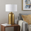 30" Gold Novelty Table Lamp With Drum Shade displayed on a dark wood tabletop in a furnished interior against a light gray wall.