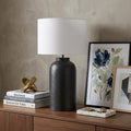 30" Black Novelty Table Lamp With Drum Shade displayed on a dark wood tabletop in a furnished interior against a tan wall.