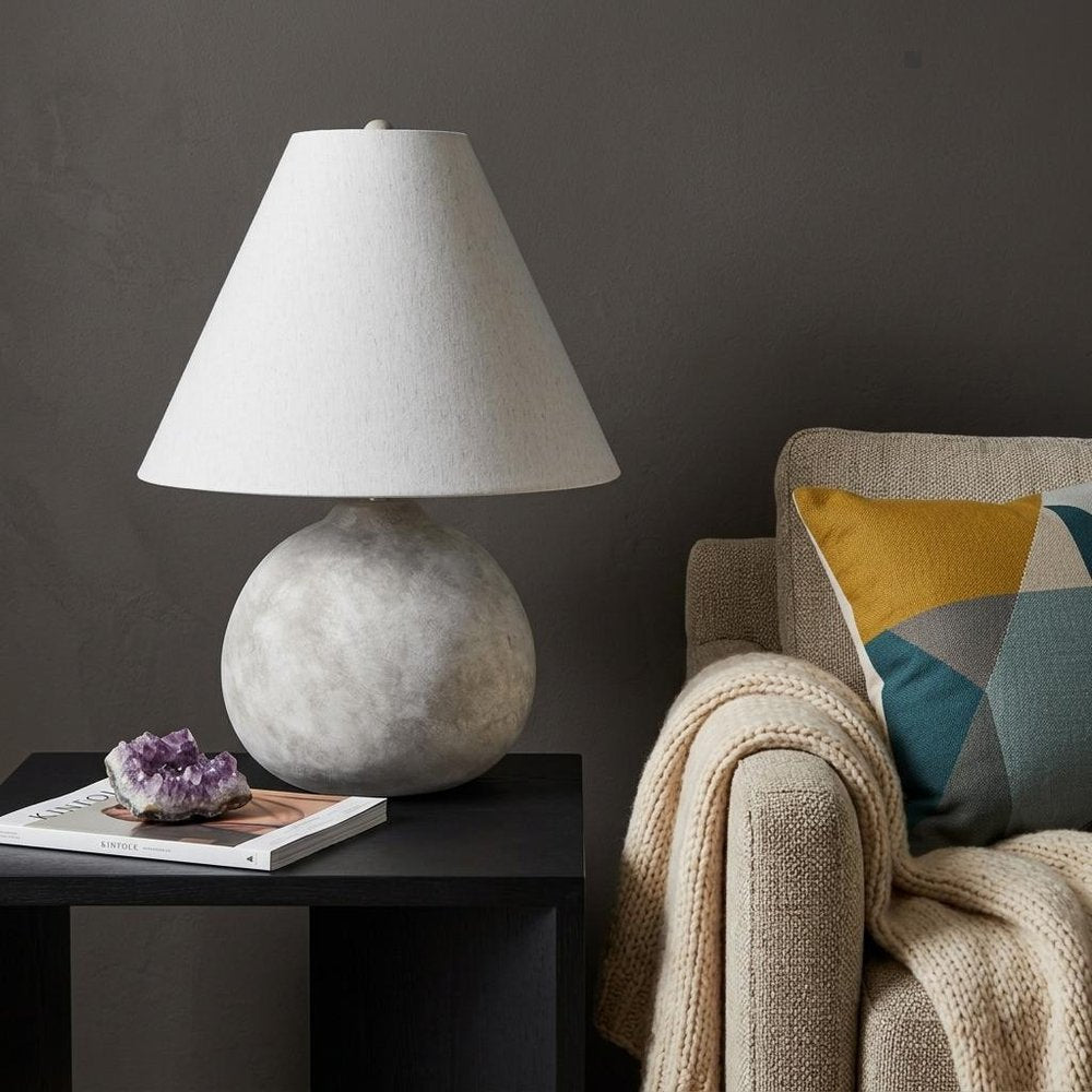 25" Tan Novelty Table Lamp With Bell Shade displayed on a tabletop in a furnished interior against a dark wall in charcoal.