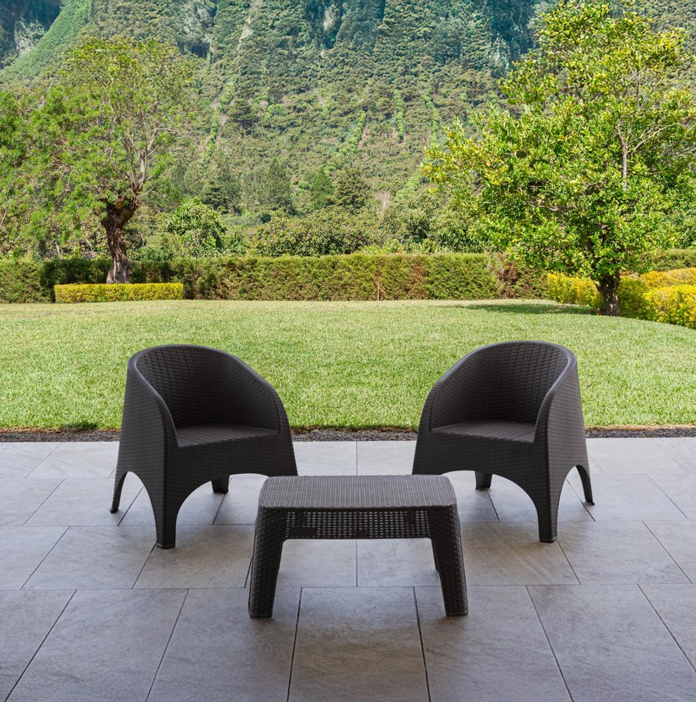 Two dark brown wicker chairs and a matching coffee table on a stone patio with a green lawn and trees in the background.