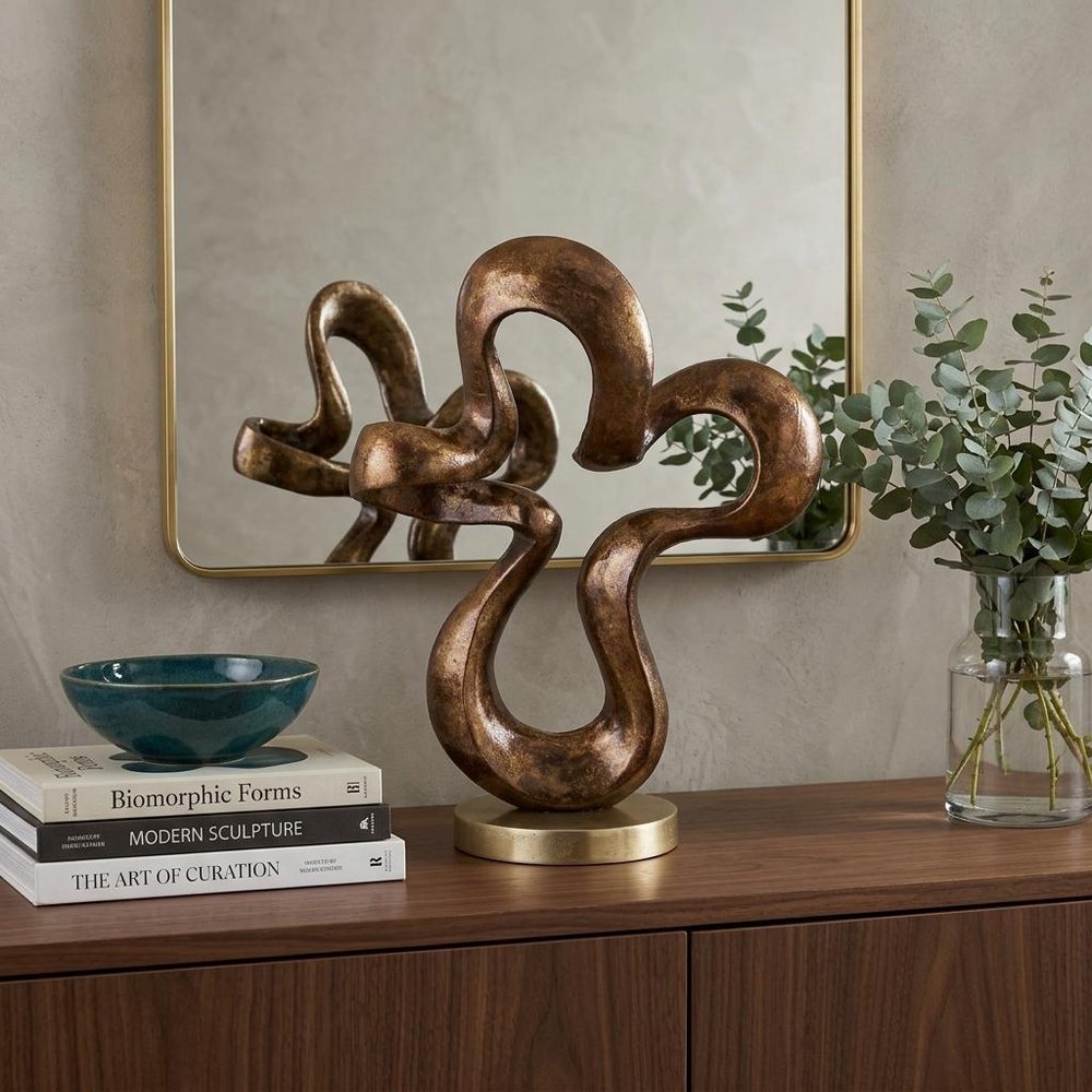 16" Bronze And Gold Aluminum Hand Painted Modern Abstract Sculpture styled in a furnished interior with a tan wall on a dark wood floor.